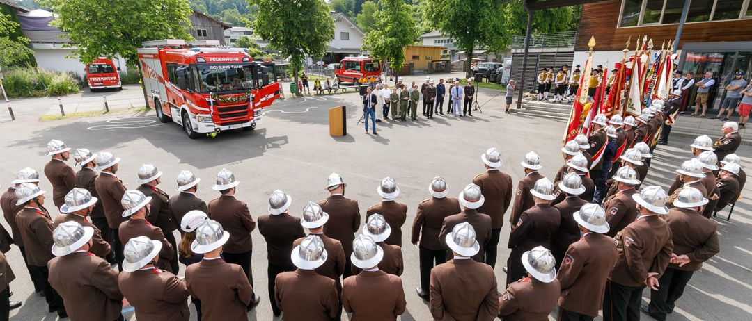 Ein roter Feuerwehrwagen steht auf einem Parkplatz mit einer Menschenmenge in braunen Uniformen. Ein Mann hält eine Rede auf einem Podium. Bäume und Häuser sind im Hintergrund.