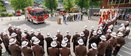 Ein roter Feuerwehrwagen steht auf einem Parkplatz mit einer Menschenmenge in braunen Uniformen. Ein Mann hält eine Rede auf einem Podium. Bäume und Häuser sind im Hintergrund.