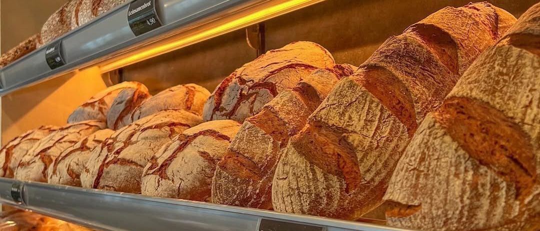 Eine Bäckerei-Vitrine mit verschiedenen Brotsorten, darunter Sonnenblumen Brot, Landbrot und anderen, beleuchtet von gelben Lichtern.
