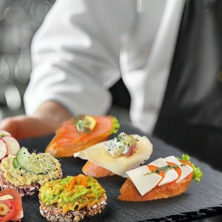 Ein Koch präsentiert eine Auswahl an Gourmet-Canapes auf einem schwarzen Tablett, darunter Räucherlachs, Avocado und Mozzarella.