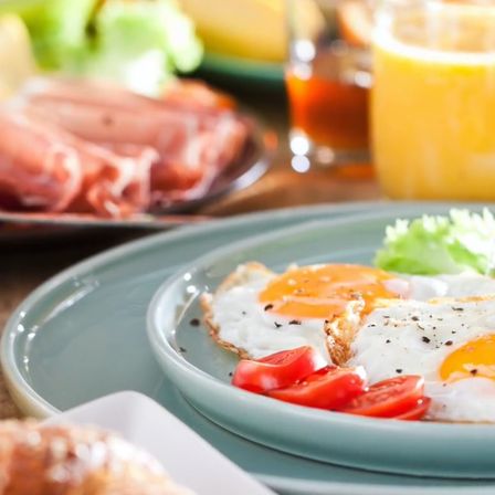 Ein Frühstücksteller mit zwei gebratenen Eiern, Tomatenscheiben und einer Seite Brot. Andere Gerichte umfassen Fleisch, Käse und ein Glas Orangensaft.