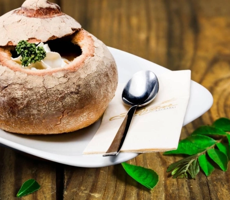 Eine Schüssel Suppe in einem ausgehöhlten Brotlaib auf einem weißen Teller mit Löffel und Serviette auf einem Holztisch mit grünen Blättern.