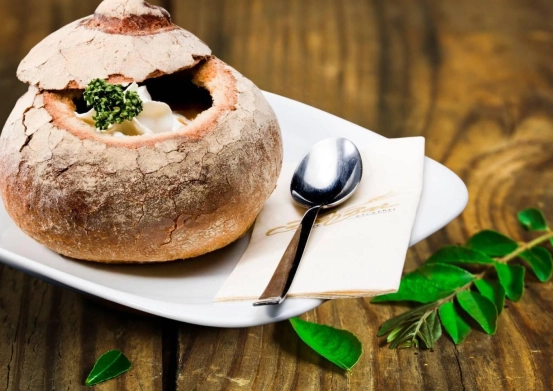 Eine Schüssel Suppe in einem ausgehöhlten Brotlaib auf einem weißen Teller mit Löffel und Serviette auf einem Holztisch mit grünen Blättern.