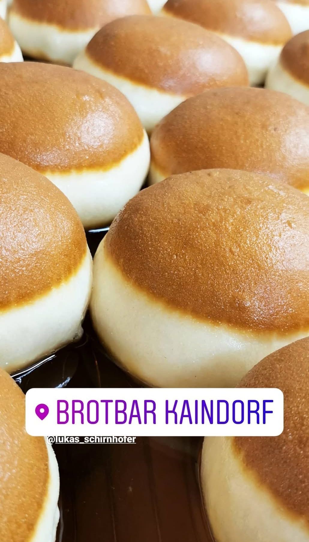 Ein Nahaufnahme von mehreren runden, goldbraunen Brötchen mit einer glänzenden, leicht glänzenden Textur. Die Brötchen sind auf einer dunklen Oberfläche präsentiert, und es gibt ein Wasserzeichen in der unteren rechten Ecke.