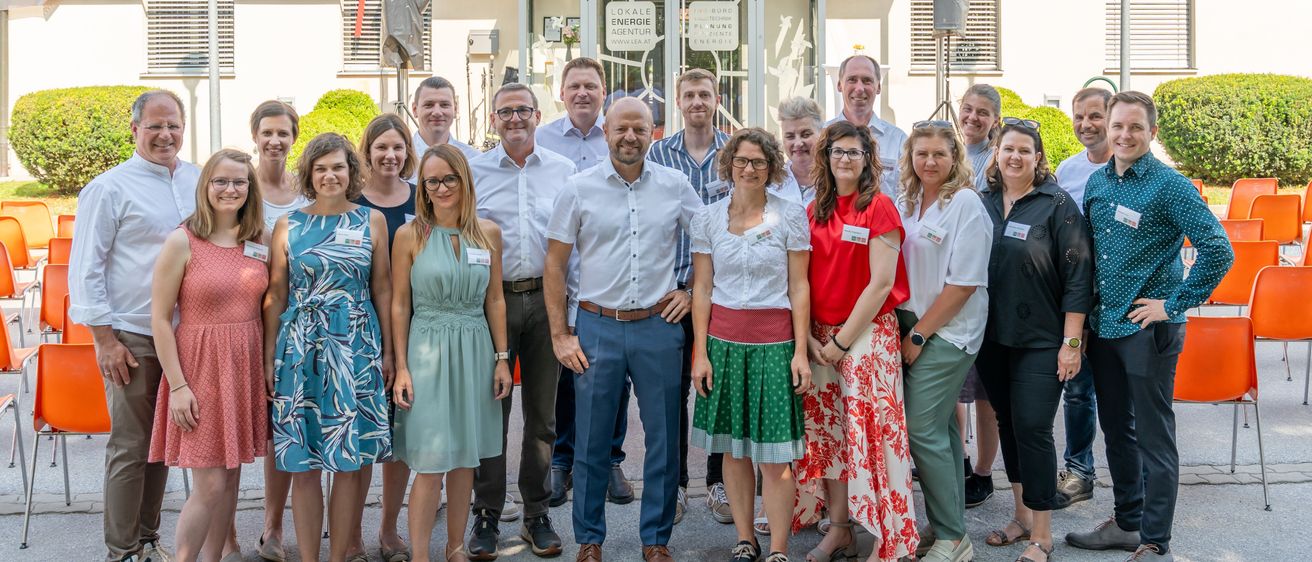 Eine Gruppe von Menschen in Sommerkleidung steht vor einem Gebäude mit dem Schriftzug 'Lokale Energieagentur'. Sie lächeln und scheinen für ein Foto zu posieren. Das Gebäude hat Glastüren und Fenster.