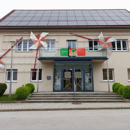 Ein Gebäude mit Solarpaneelen auf dem Dach und Windrädern über dem Eingang, mit einer verglasten Terrasse und einem Schild, das 'Energie Agentur' besagt.