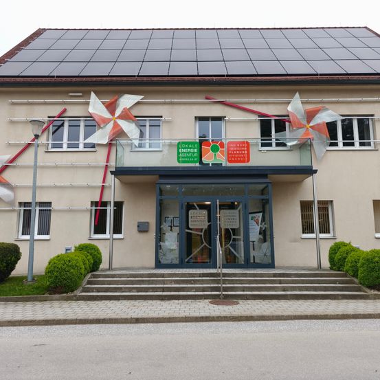 Ein Gebäude mit Solarpaneelen auf dem Dach und Windrädern über dem Eingang, mit einer verglasten Terrasse und einem Schild, das 'Energie Agentur' besagt.
