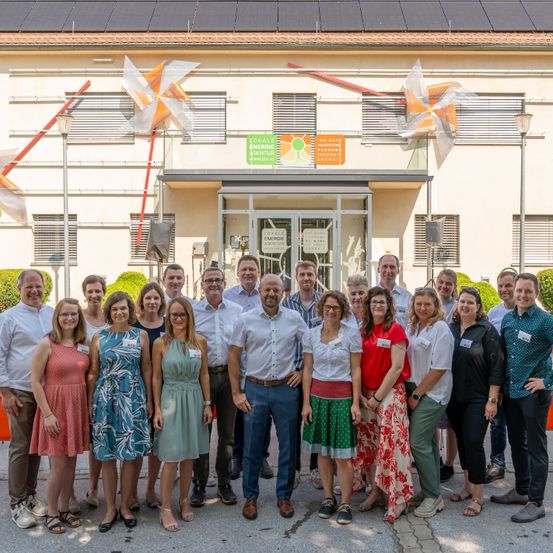 Eine Gruppe von Menschen steht vor einem Gebäude mit Solarpanelen und Windrädern. Sie sind in Sommerkleidung gekleidet, einige mit Brillen, und scheinen für ein Foto zu posieren.