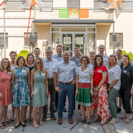 Eine Gruppe von Menschen steht vor einem Gebäude und lächelt für ein Foto. Das Gebäude hat Schilder wie 'lokalenergie' und 'inge'. Einige tragen Brillen, und es gibt Flaggen und Pflanzen in der Umgebung.