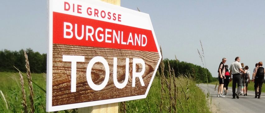Ein Schild für 'Die Grosse Burgenland Tour' ist an einem Holzpfahl am Straßenrand befestigt, mit einer Gruppe von Menschen, die im Hintergrund auf der Straße gehen.