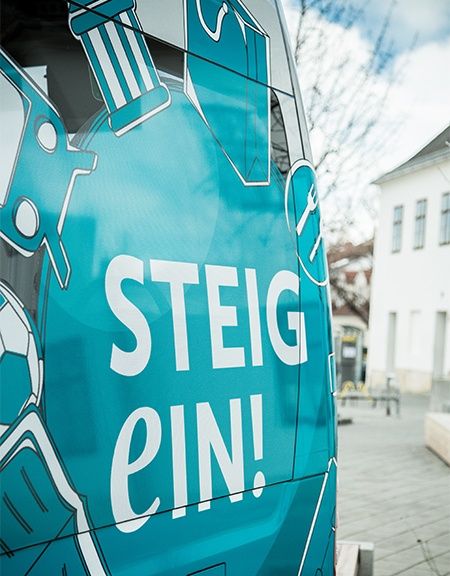 Ein blaues Fahrzeug mit einer weißen Illustration eines Fußballs, eines Bleistifts und eines Buches. Die Worte STEIG EIN! sind in Weiß geschrieben. Im Hintergrund befindet sich ein Gebäude mit Glasfenstern und einem Baum.