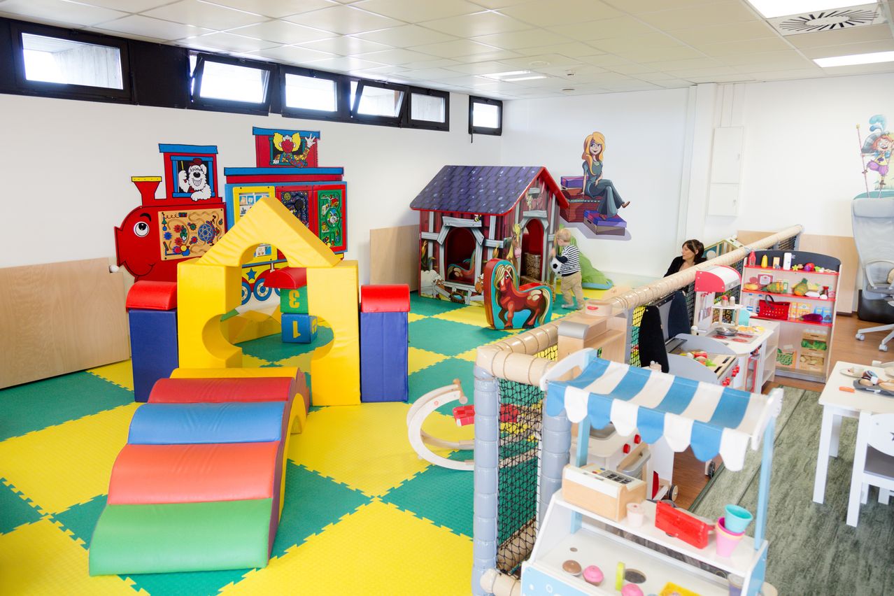 Ein farbenfroher Indoor-Spielbereich mit verschiedenen Spielzeugen und einem Spielhaus. Kinder spielen, und eine Frau sitzt in der Nähe. Eine Cartoon-Figur ist an der Wand gemalt.