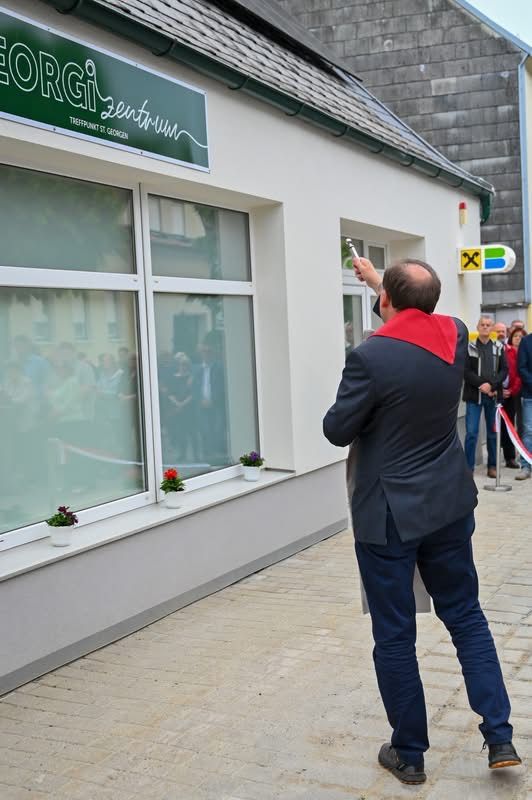 Ein Mann schneidet das Band eines Gebäudes namens Georg. Mehrere Leute schauen zu. Es gibt Blumen in Töpfen auf der Fensterbank.