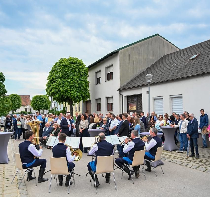 Eine Band spielt verschiedene Instrumente vor einem Gebäude, das von vielen Menschen beobachtet wird. Die Musiker sitzen auf Stühlen, und im Hintergrund befinden sich Bäume und eine Straßenlaterne.