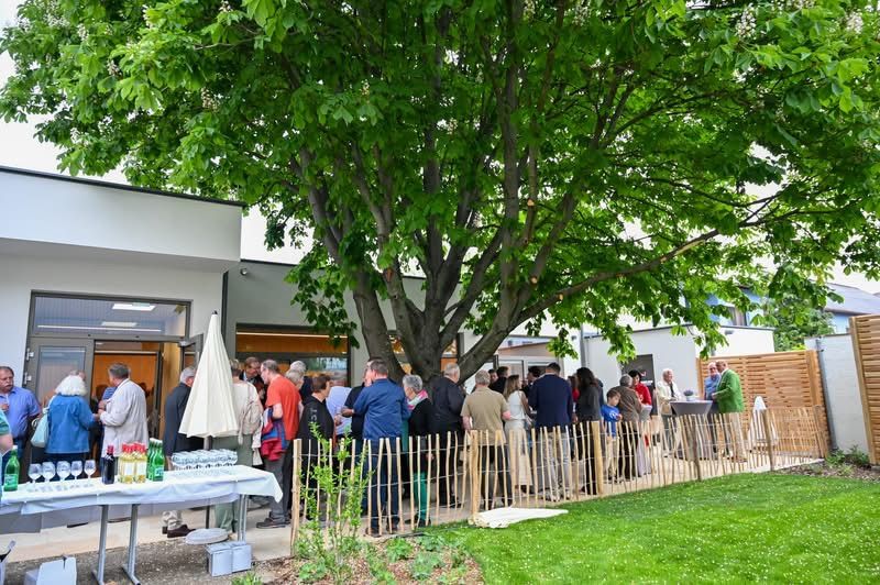 Menschen versammeln sich unter einem Baum bei einer Veranstaltung. Einige stehen in der Nähe eines Tisches mit Weingläsern, während andere in der Schlange am Eingang warten.