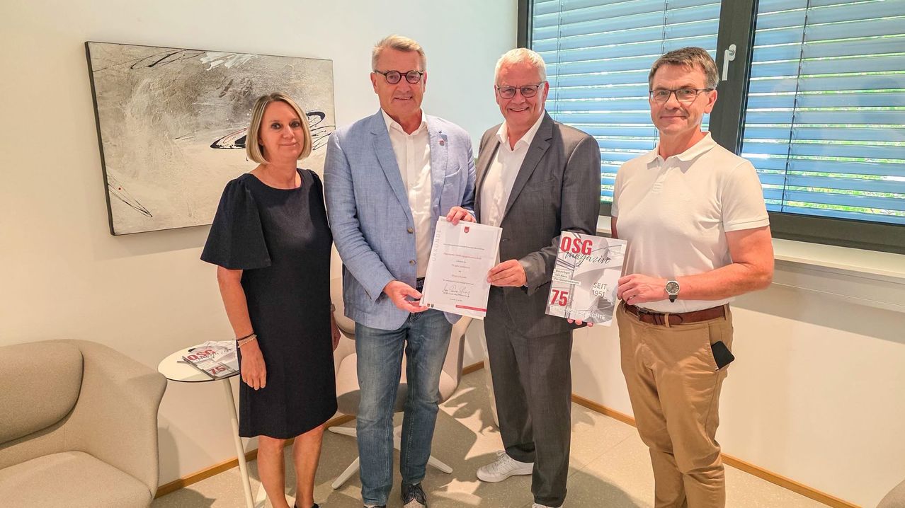 Vier Personen stehen zusammen in einem Raum, lächeln und posieren für ein Foto. Der Mann in der Mitte hält ein Zertifikat und eine Zeitschrift. Der Mann auf der rechten Seite hält eine Zeitschrift. Die Frau auf der linken Seite trägt eine Armband. Der Raum hat einen weißen Tisch und ein Gemälde an der Wand.