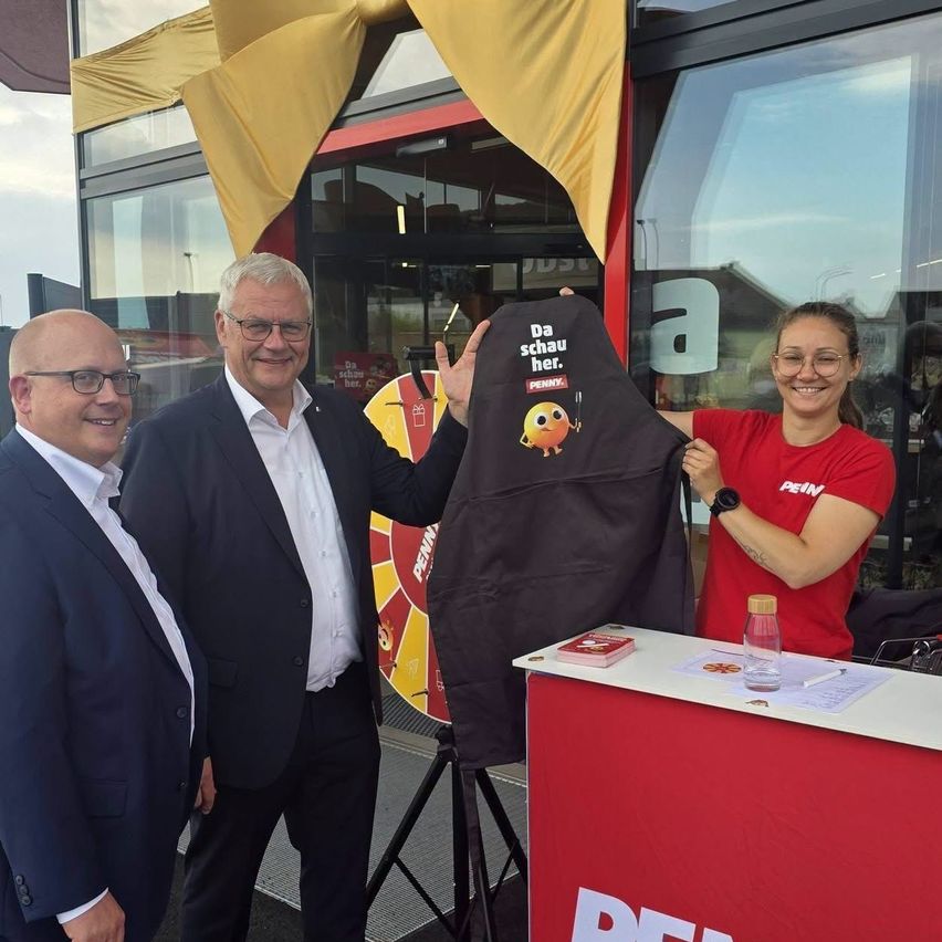 Drei Männer in formeller Kleidung stehen vor einem Geschäft, einer hält eine schwarze Schürze mit einem gelben Logo und Text. Eine Frau in einem roten Shirt hält die Schürze hoch.