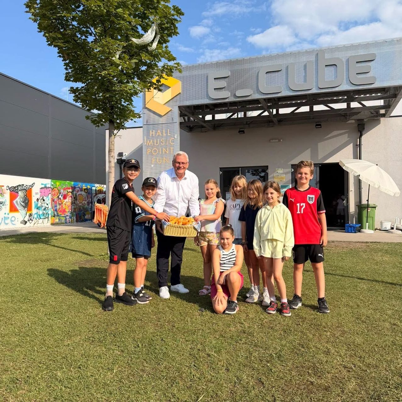 A group of children and adults stand in front of a building with a sign that reads E.cube. The man in the middle holds a basket of food.