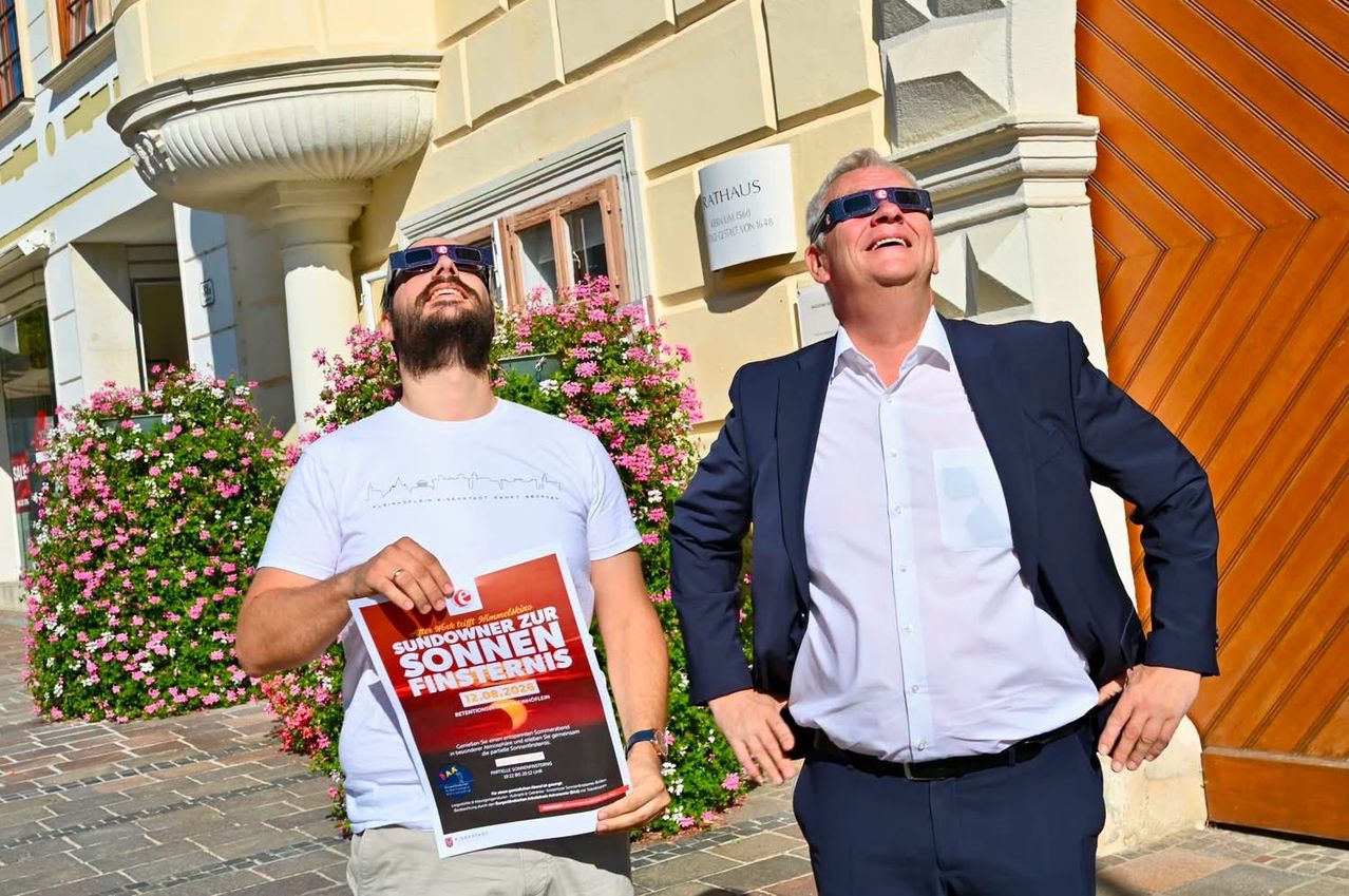 Two men, one in a white shirt and the other in a blue suit, stand outside a building with a sign that reads 'SUNOWNER FOR SONNEN FINSTERNIS'. The man in white holds a poster with the same text and a date, while the man in blue looks up at the sky.