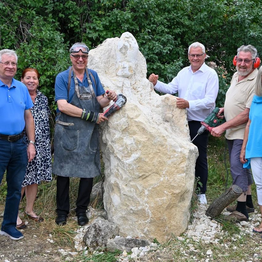 A group of people stand around a large stone sculpture, two men hold drills, one wears goggles, and another wears headphones. They are all smiling.