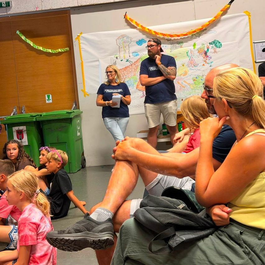 A group of children and adults are sitting on the floor in a room, listening to a man and woman standing in front of a large drawing on a wall.