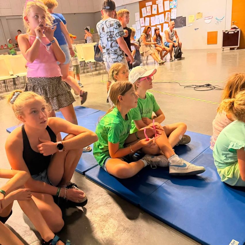 A group of children and young adults sit on blue mats in a room, some wearing bracelets and sneakers. A few adults stand and sit in the background.