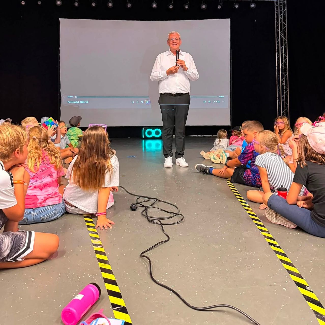 A man in a white shirt and glasses stands on a stage with a microphone, addressing a group of children sitting on the floor in front of him. The stage has a large screen behind him, and there are yellow and black caution tape lines on the floor.