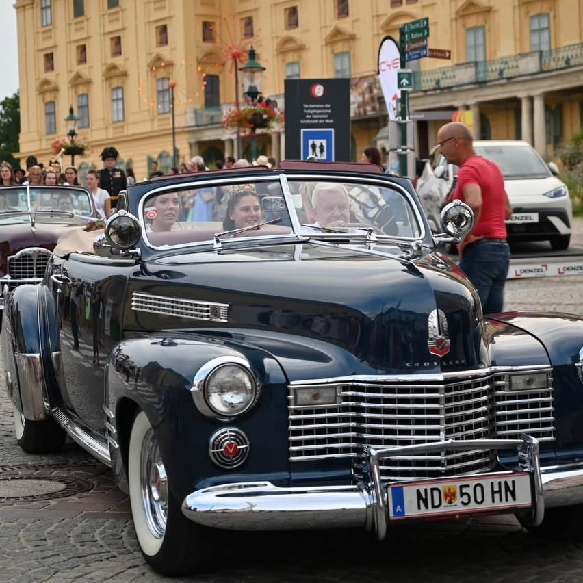 A vintage blue Cadillac with a German license plate ND 50 HN is parked on a cobblestone street. A man and two women are sitting inside. Behind the car, a crowd of people is gathered, and a building with many windows is in the background.