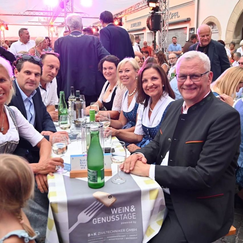 A group of people are sitting at a table with wine glasses, bottles, and a tablecloth that reads 'Wein & Genussstage.' They are smiling and appear to be enjoying a meal or drinks. The setting looks like a festive event.