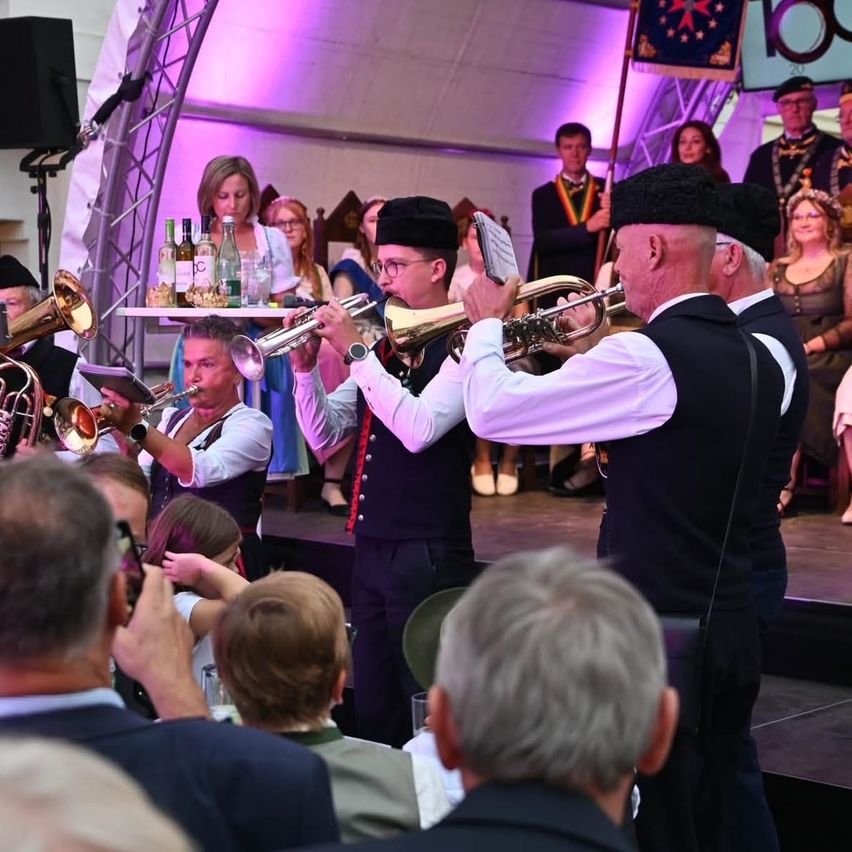 A group of musicians plays trumpets on stage at an event, with a crowd watching in front. The stage is decorated with bottles and a flag.