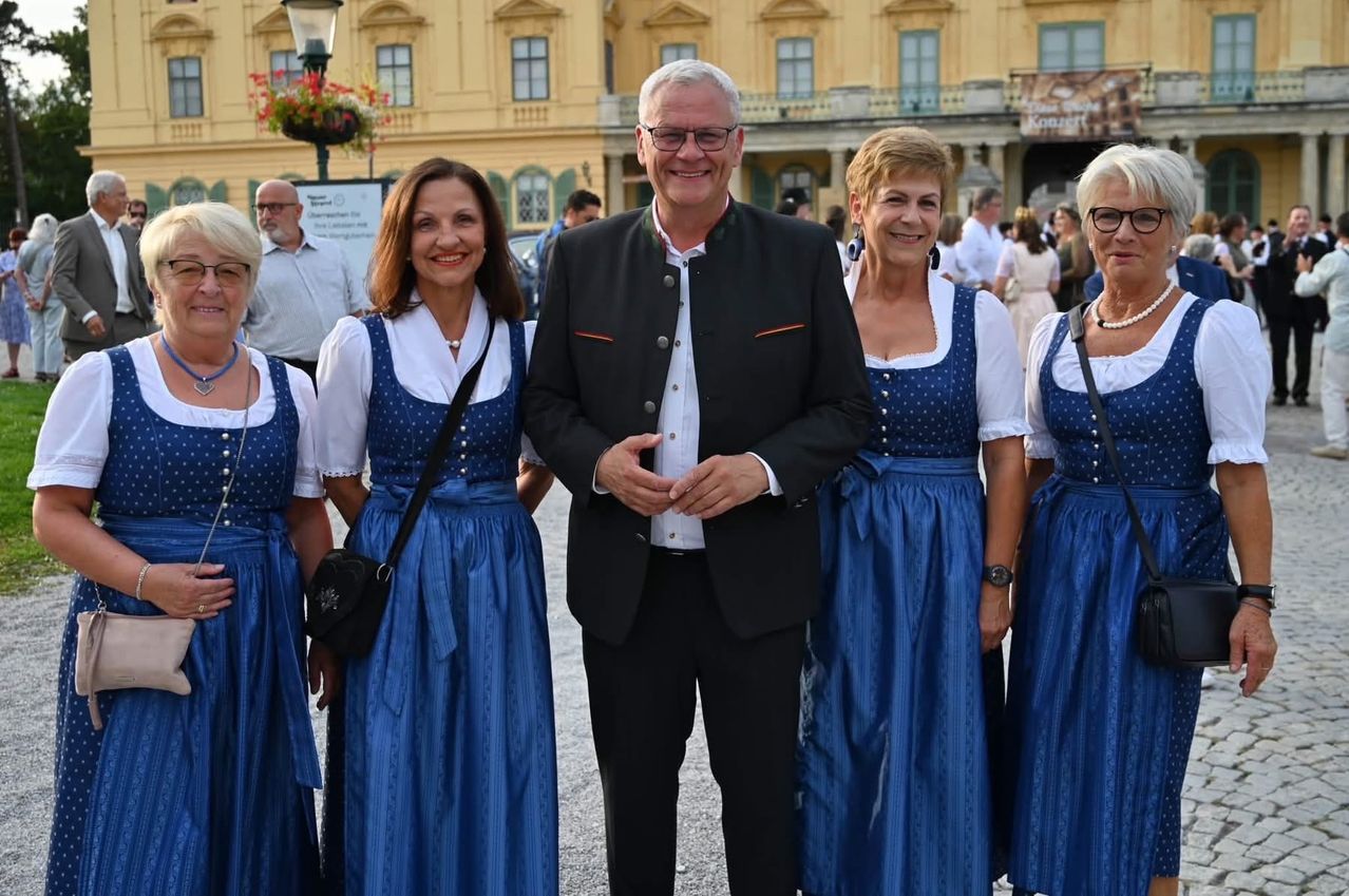 A man in a black suit stands between four women in blue dresses in front of a large building.