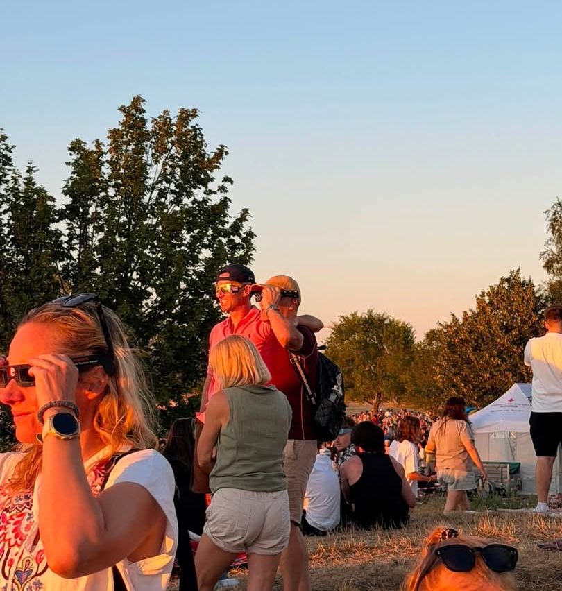 A group of people at an outdoor event, some wearing sunglasses and hats, with trees and a tent in the background.