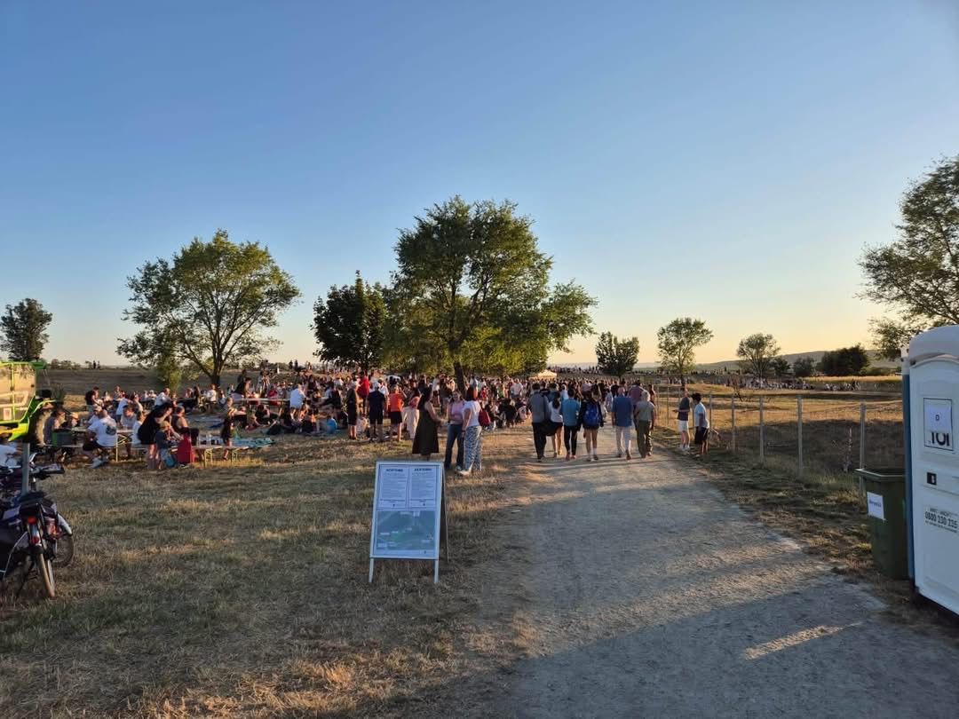 A large crowd of people are gathered in an open field, some sitting on the grass while others walk along a dirt path. Trees and a fence are visible in the background.