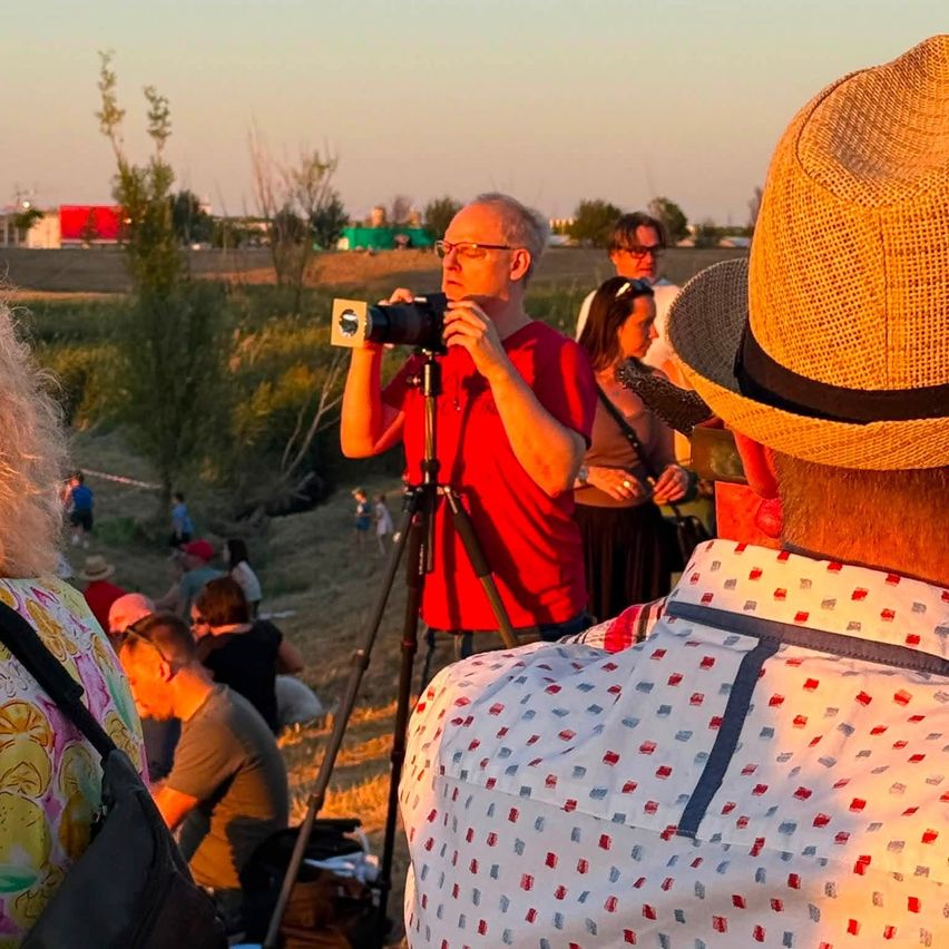 A group of people are gathered outdoors, some are sitting while others are standing. A man in a red shirt is taking a photo with a camera on a tripod.