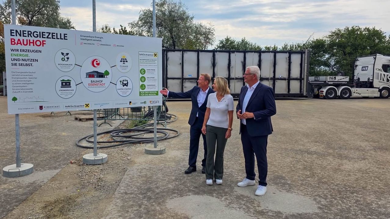 Three individuals, two men and a woman, stand in front of a large signboard, possibly at a construction site. They are dressed formally, and the man on the left is gesturing towards the sign. Behind them, there is a large, white structure with a metal fence and some trees.