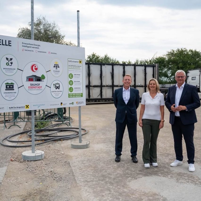 Three individuals stand in front of a large informational sign, likely at a renewable energy facility. The sign includes diagrams and text about biogas, solar power, and waste-to-energy.