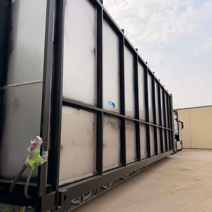A large, black storage container with multiple glass panels and a small object hanging from one corner. The container is parked in an outdoor area.