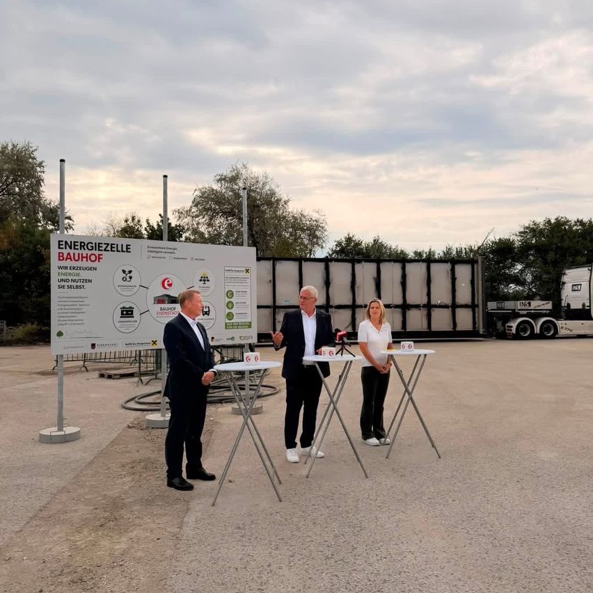 Three individuals stand before a large sign in front of a building, discussing with microphones in a parking lot.
