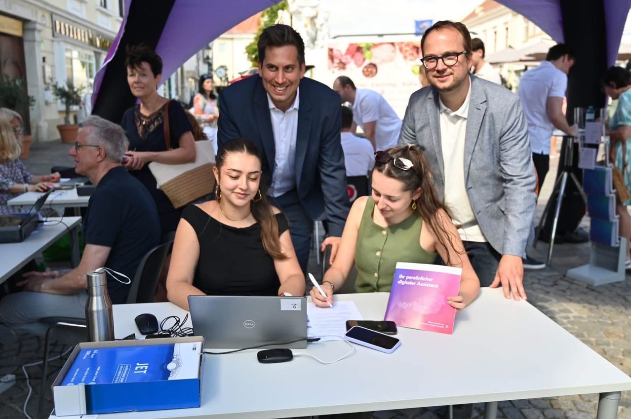 Two women sit at a table with a laptop, a book, and a pen. Behind them, two men stand and smile. They appear to be at an outdoor event.