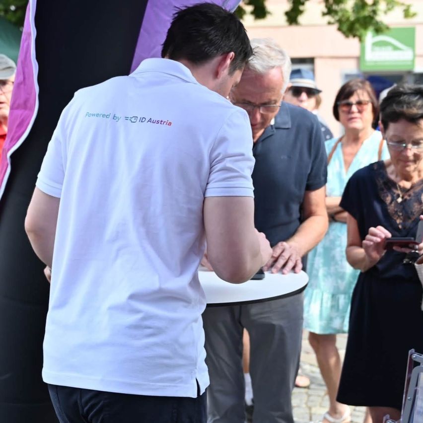 A man in a white shirt with 'Powered by ID Austria' written on the back stands at a table, facing a group of people, one of whom is using a smartphone.