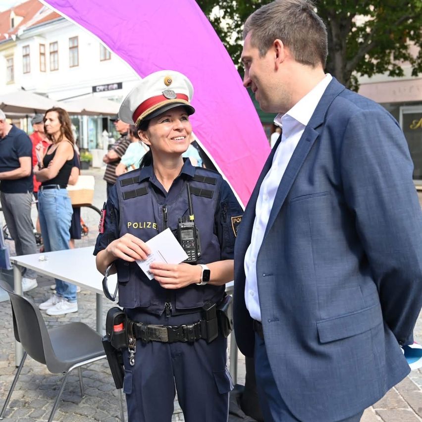 A police officer in uniform is speaking with a man in a suit. They are standing in front of a table with a purple banner behind them.