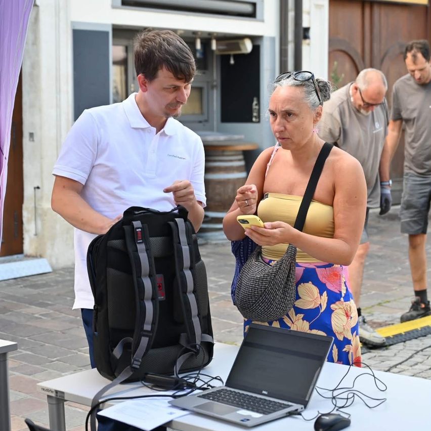 A woman in a yellow top and floral skirt looks at her phone while a man in a white shirt stands beside her with a backpack and laptop on a table.