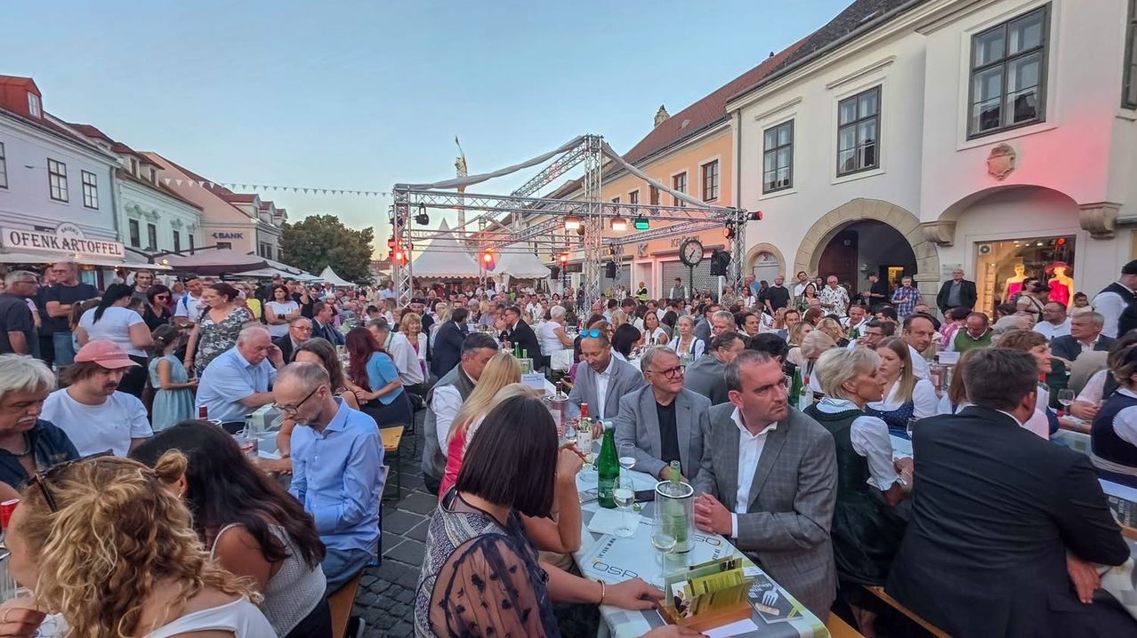 Eine große Menschenmenge versammelt sich in einer Freiluftveranstaltung, möglicherweise für ein Festival, mit aufgestellten Tischen, Stühlen und Zelten. Das Gebiet ist von Gebäuden umgeben, und im Hintergrund befinden sich Straßenlaternen und eine Bühne.