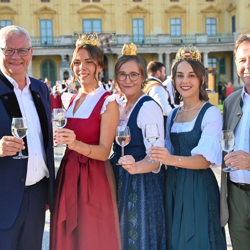 Drei Frauen in traditionellen Kleidern und goldenen Kronen, zusammen mit zwei Männern, halten Weinglaser vor einem historischen Gebäude.