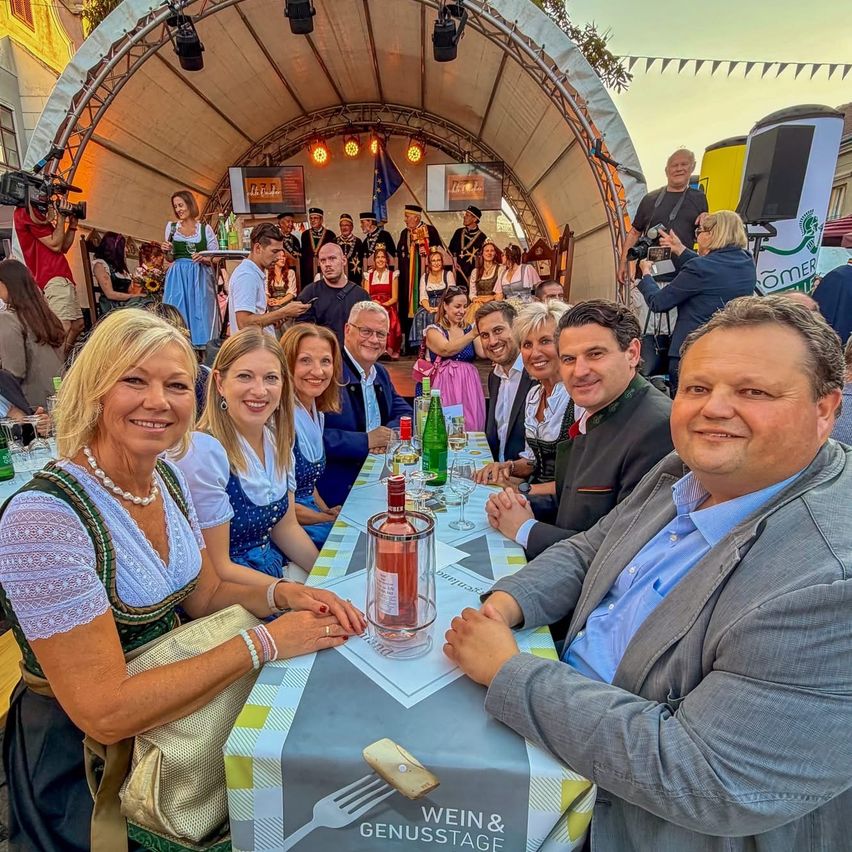 Eine Gruppe von Menschen, darunter Männer und Frauen, sitzen an einem Tisch mit Weinflaschen und Gläsern, lächeln und posieren für ein Foto auf einem Festival.