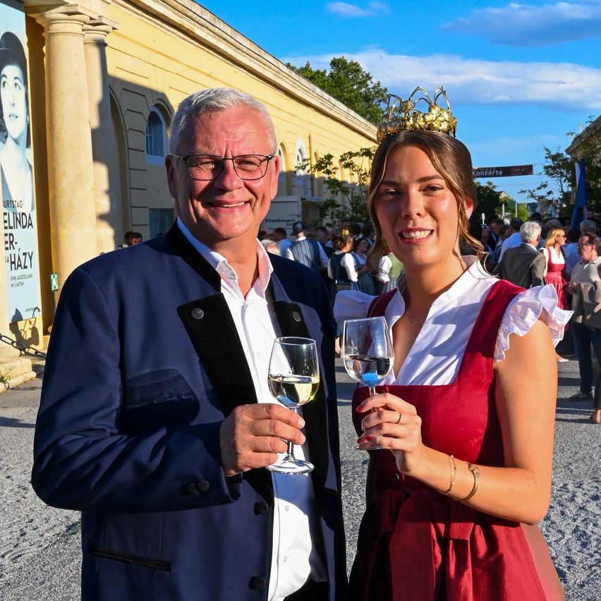 Ein Mann und eine Frau in traditioneller Kleidung halten Weinglas vor einem historischen Gebäude.
