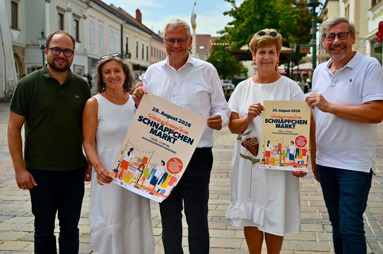 Vier Personen stehen auf einer Kopfsteinpflasterstraße und lächeln. Zwei Frauen halten Schilder mit der Aufschrift 'Schnäppchenmarkt'. Der Mann in der Mitte hält ein größeres Schild. Hinter ihnen befinden sich Gebäude und Bäume.