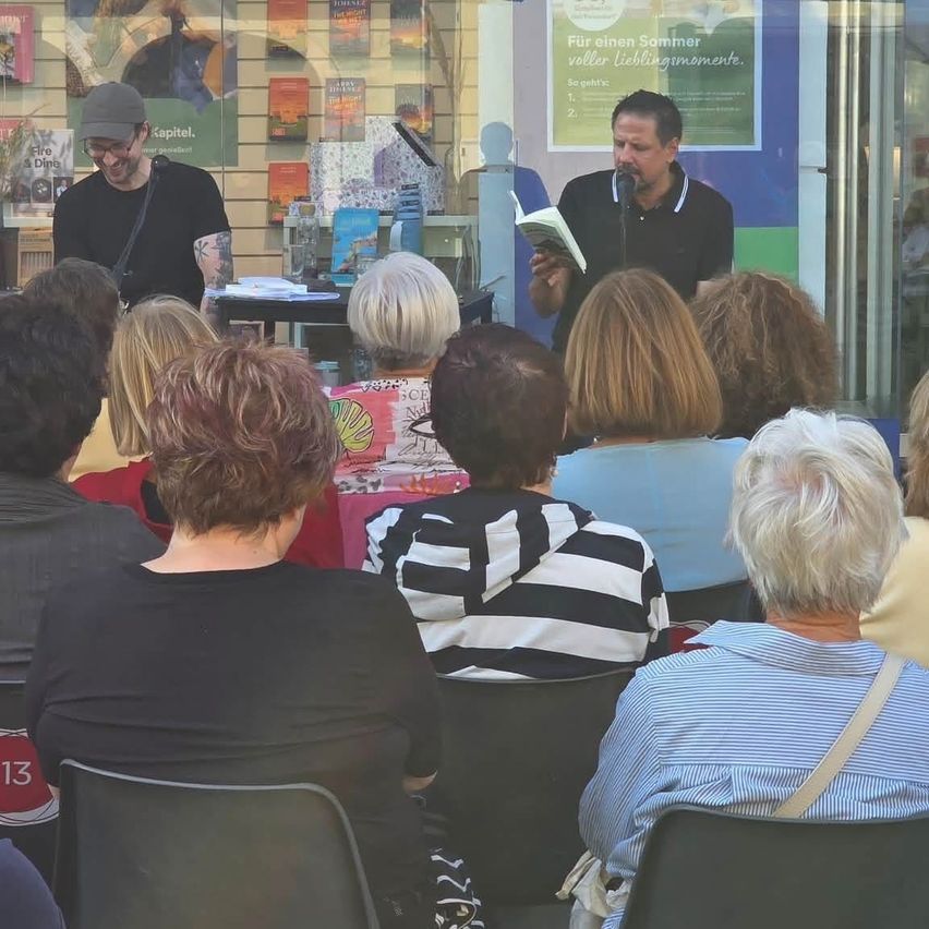 Ein Mann liest ein Buch in ein Mikrofon, während eine Gruppe von Menschen in Stühlen vor ihm sitzen. Ein anderer Mann steht hinter ihm mit einer Tasche.