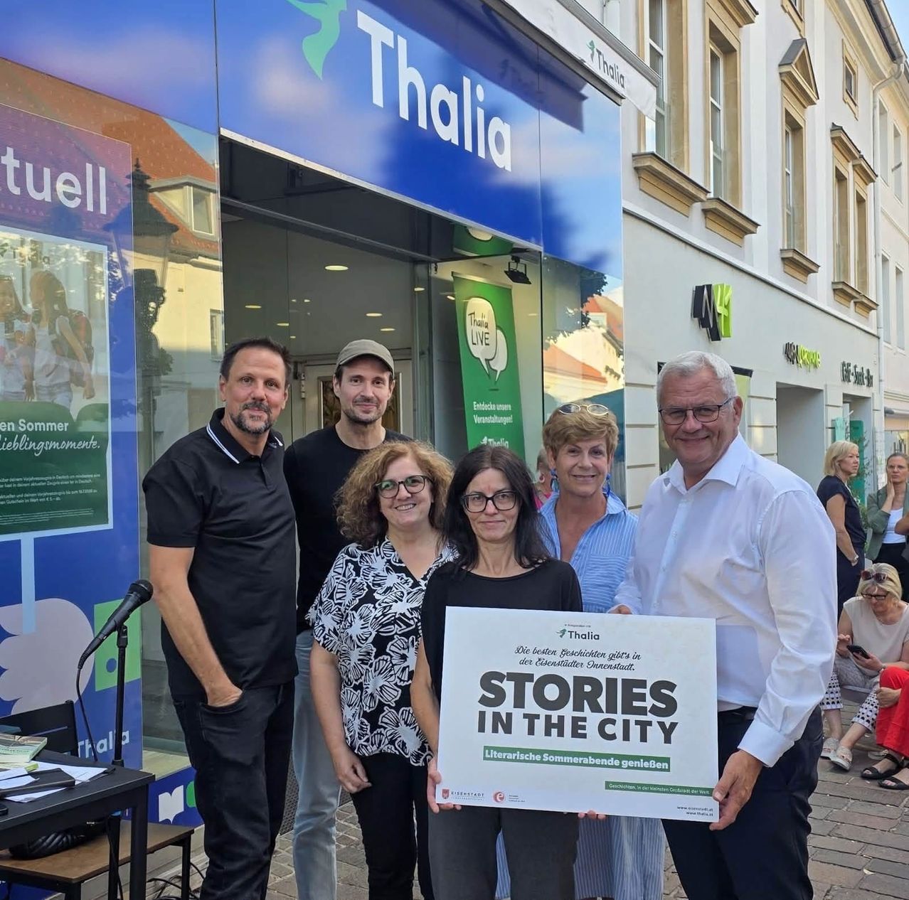 Fünf Personen stehen vor einem Thalia-Buchladen und halten ein Schild mit der Aufschrift 'Stories in the City'. Dahinter ist ein Gebäude mit einem Schild zu sehen, das 'Thalia' besagt.