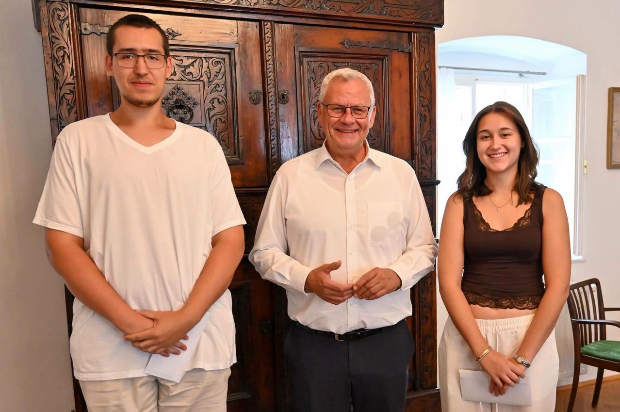 Drei Personen stehen gemeinsam vor einem Holzschrank. Der Mann links trägt eine Brille und ein weißes T-Shirt, der Mann in der Mitte trägt ein weißes Hemd und eine Brille, und die Frau rechts trägt ein braunes Oberteil und weiße Hose.